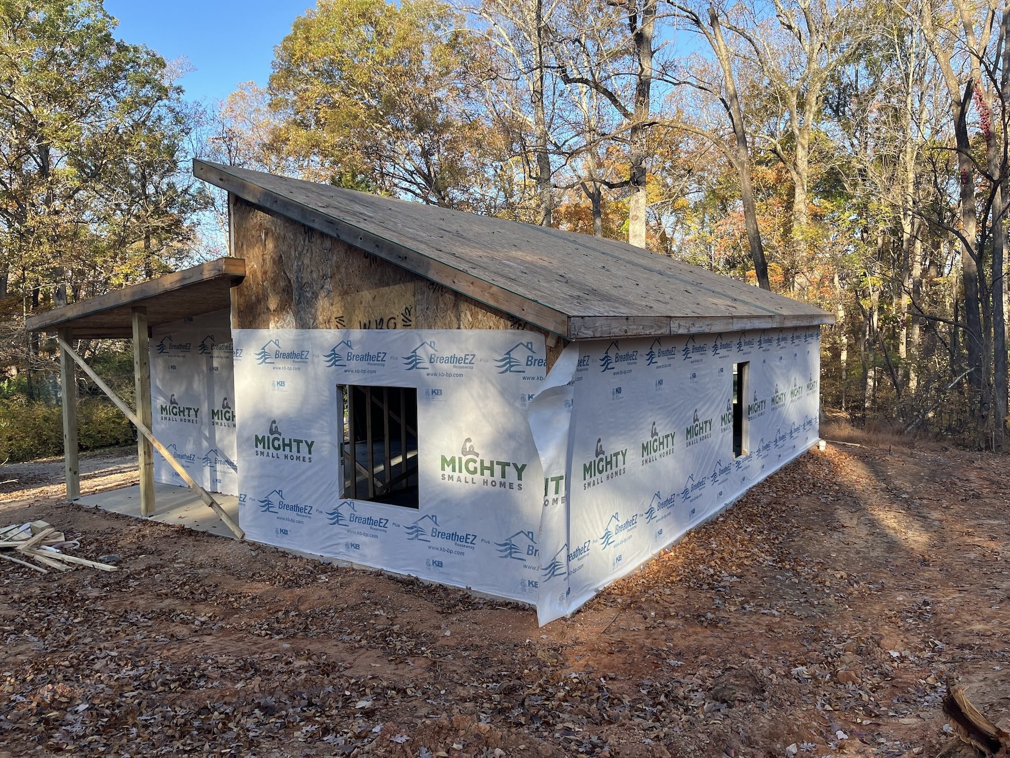 A partially finished tiny house with a wooden frame, built by Mighty Small Homes, set in a wooded area with exposed dirt and construction materials around.