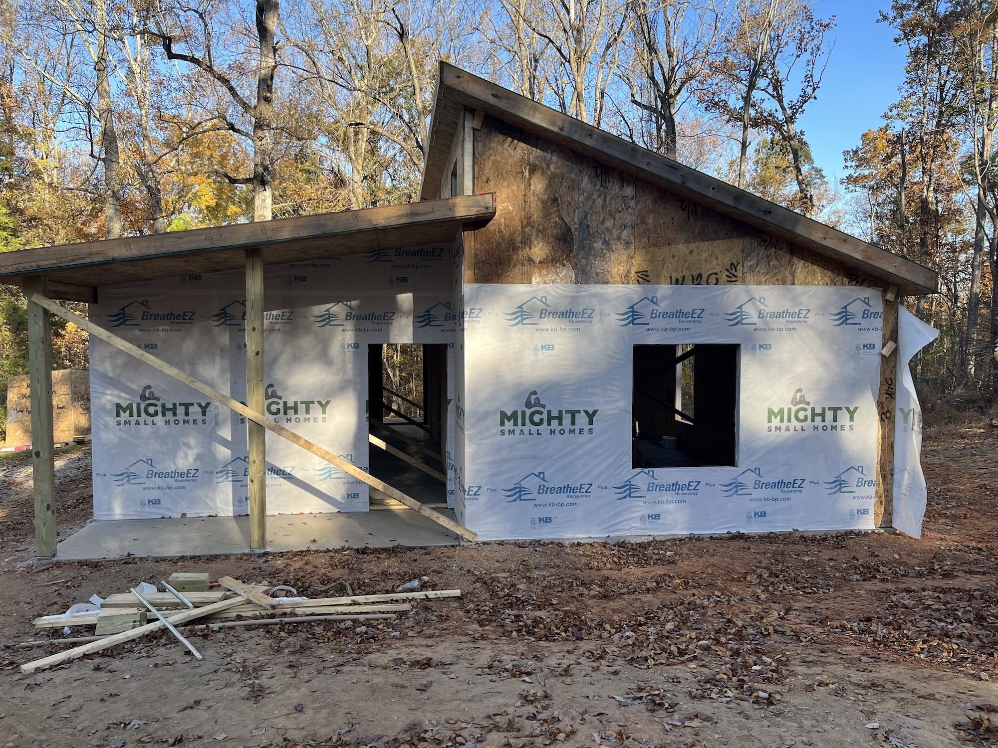 Side view of a small modern building with wooden walls covered in house wrap with the Mighty Small Homes logo and a sloped roof sitting in a wooded area. The side of the building has openings for a door and a single window. The trees are bare, suggesting early spring or late fall.