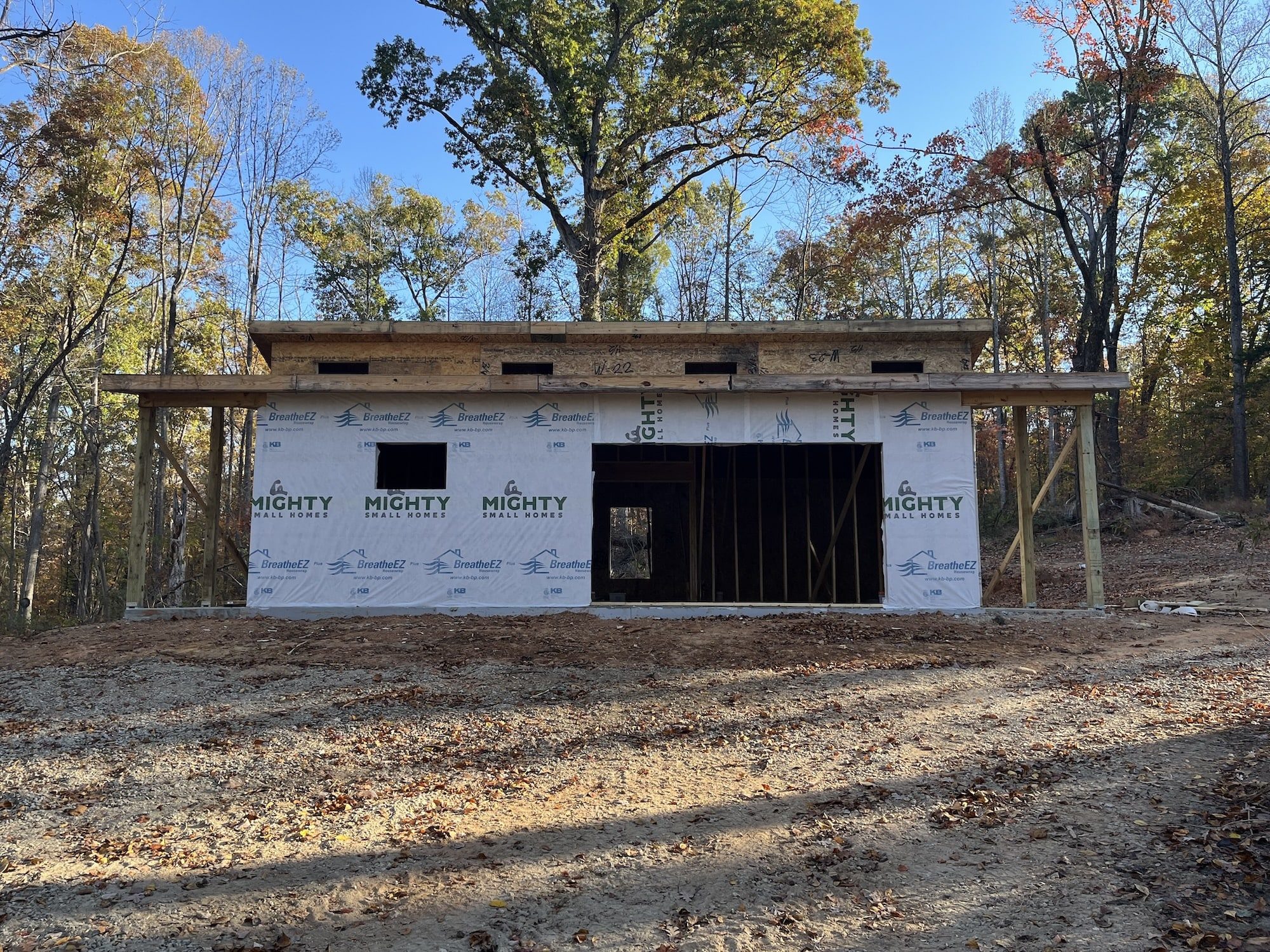 A small modern building with a wooden frame and house wrap with the Mighty Small Homes logo sits in a wooded area. The building has a large opening for sliding glass doors and one for a single window. The trees are bare, suggesting early spring or late fall.