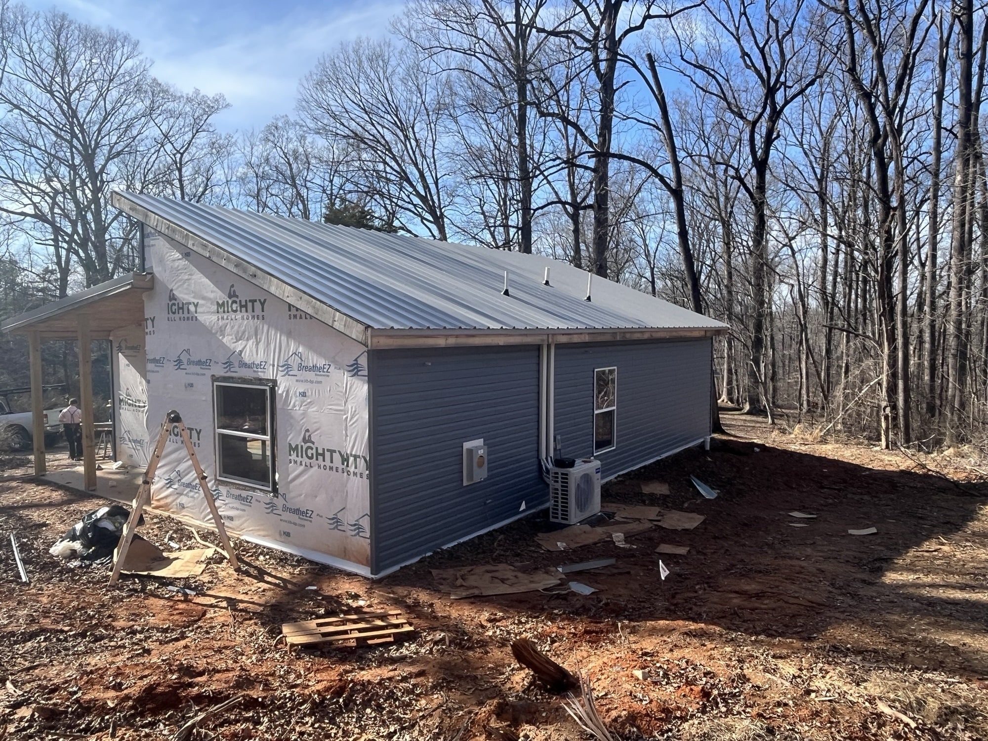A partially finished tiny house with gray metal roof and siding, built by Mighty Small Homes, set in a wooded area with exposed dirt and construction materials around.