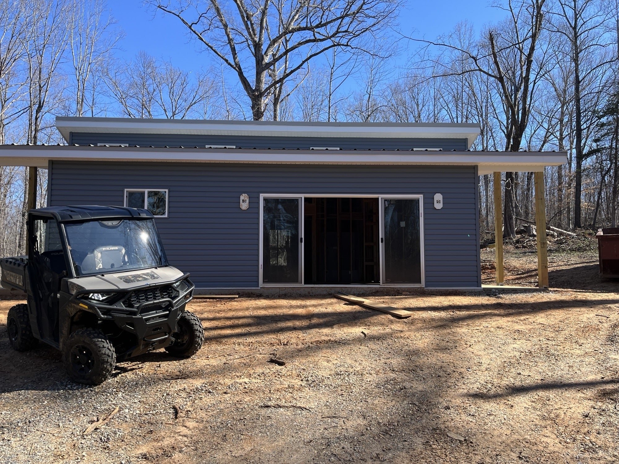 A small modern building with blue siding and a sloped roof sits in a wooded area. A black utility vehicle is parked in front, and the building has large sliding glass doors and a single window. The trees are bare, suggesting early spring or late fall.