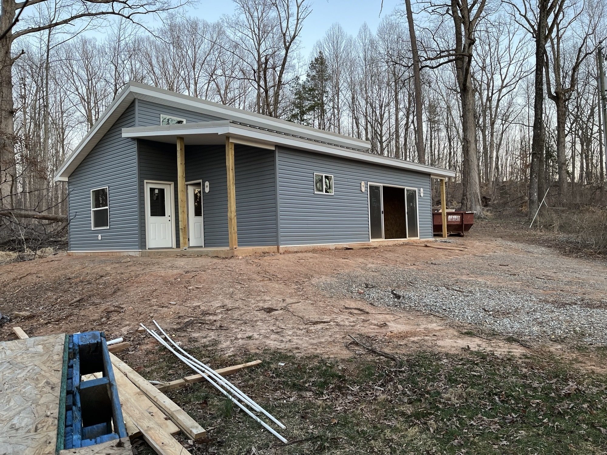 A nearly completed modern tiny house by Mighty Small Homes, featuring blue-gray siding, a front porch with wooden posts, and sliding doors on the side, surrounded by trees and a gravel driveway.
