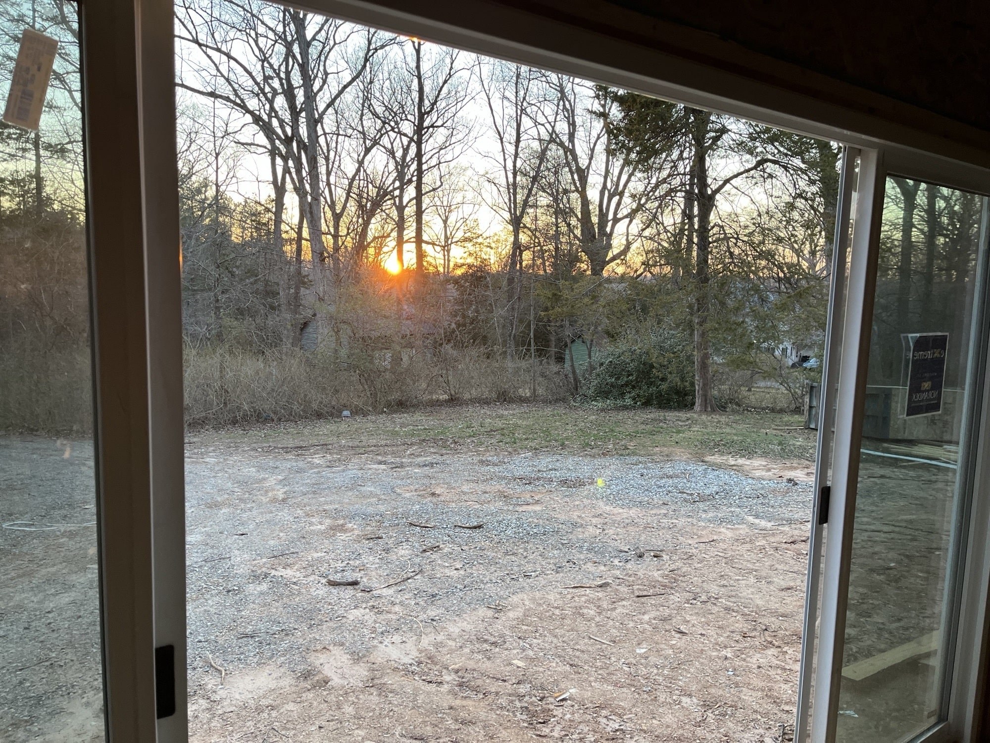 A sunset view from inside the tiny house looking out through large sliding glass doors onto a gravel-covered clearing and tree-lined surroundings.