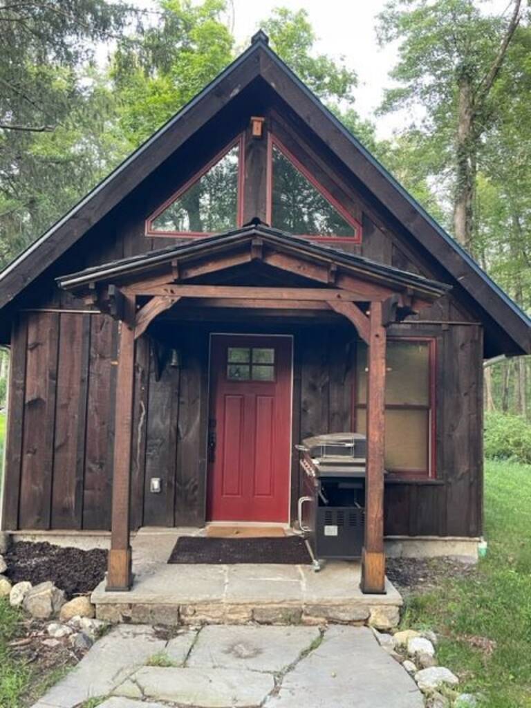 cottage model with a covered timber porch, red front door, and loft window overlooking a wooded setting, illustrating a comfortable small home suited for off-grid and rural living