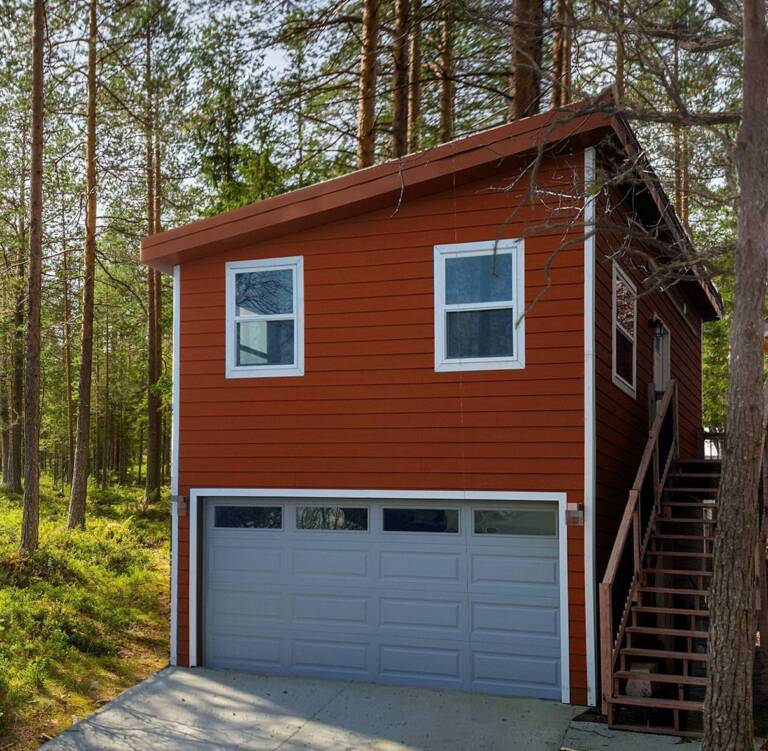 carriage model with an upper living space above a two-car garage, surrounded by trees and designed to provide flexible off-grid living, guest accommodations, storage, or workspace