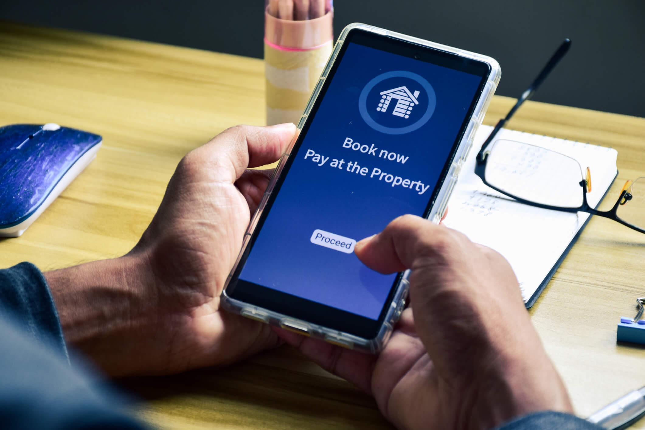 person holding a smartphone displaying a booking screen with a “Book now, pay at the property” option