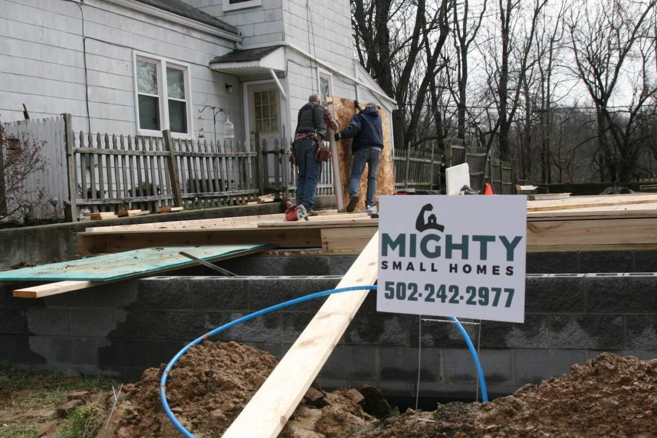 Photo of workers moving a wall panel with a Mighty Small Homes yard sign in the foreground
